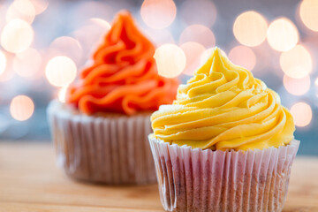 Two cupcakes decorated with orange and yellow cream and festive lights in the background.