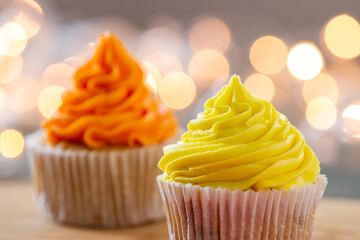 Close up of two cupcakes decorated with orange and yellow cream and festive lights in the...