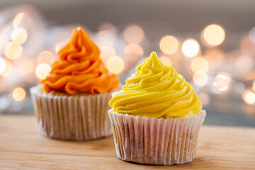 Two cupcakes decorated with orange and yellow cream and festive lights in the background.