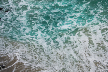 Textured seascape of breaking wave in the beach. Aerial view of turquoise water with foam reaching the sand. Travel and vacations in nature concept.