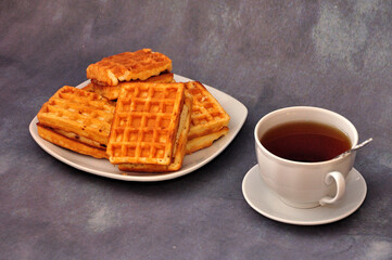 A plate with a stack of fresh Belgian waffles and a cup of hot tea on a gray background.