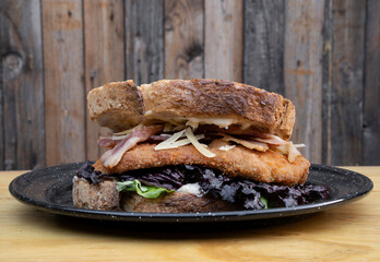 Delicious sandwich made with bread, parmesan cheese, lettuce, bacon and fried chicken steak, in a black dish on the restaurant wooden table.