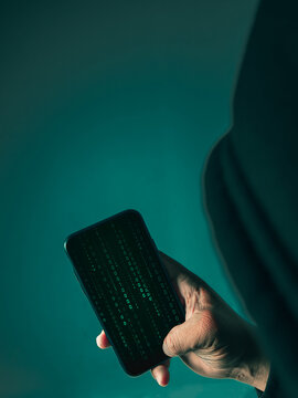 An anonymous masked hacker is using a smartphone to penetrate credit card financial information. Hacking and malware concept.