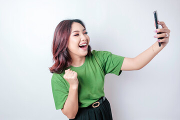 A young Asian woman with a happy successful expression wearing green t-shirt and holding smartphone isolated by white background