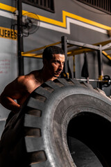 Obraz na płótnie Canvas High quality photo. Muscular shirtless Hispanic man lifting a big tire in a gym training with all his might.