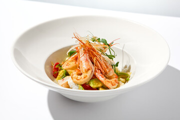 Elegant food - salad with shrimp and avocado on white table with harsh shadow. Flashy food concept. Seafood salad with shrimp, tomatoes and avocado on white plate with sunlight.