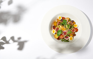 Traditional french salad nicoise with tuna, potatoes, eggs and vegetables on white table with harsh shadows. Tuna salad with quail eggs on white background with shadows of leaves.