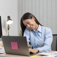 Asian female employee talking on the phone and working in a laptop with a smiling face, Office staff work and telephone conversations.