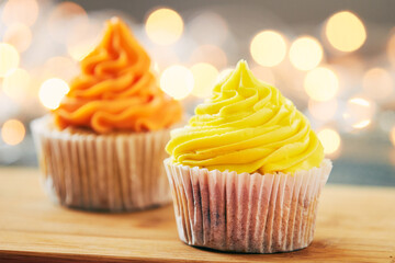 Two cupcakes decorated with orange and yellow cream and festive lights on the background.