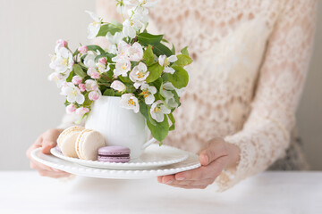 Beautiful composition with delicious French macarons and spring flowers in a white cup. Young beautiful girl in a lace dress holding a plate with sweet dessert and apple tree flower. Bride's morning