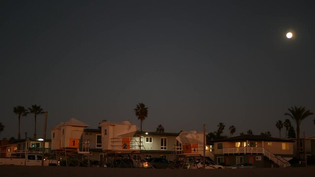 Palm trees silhouettes and full moon in twilight sky, California beach, USA. Beachfront houses or homes on coast in evening, fullmoon on pacific ocean shore in dusk. Lifeguard tower. Light in windows.