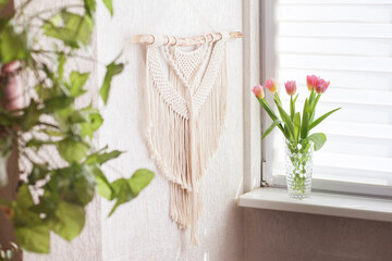 Handmade macrame wallhanging. 100% cotton wall decoration with wooden stick hanging on a white wall.    Female hobby.  ECO friendly modern concept in the interior.