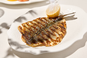Grilled dorado fish on light background with shadows. Grilled dorado with lemon on white table. Aesthetic composition with bbq fish. Bbq dorado. Grilled fish. Summer menu.