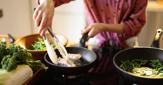 Woman turning salmon steaks on pan and mixing salad, cooking healthy mediterranean food in the kitchen at home, close-up with no face