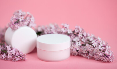 Face and body skin care. A set of cosmetic creams and balms in white tubes and cans on a coral background with sprigs of lilac flowers. Spa treatments for home care. Home rejuvenation and moisturizing