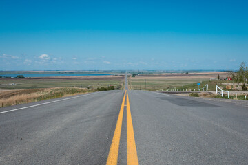 road with two yellow lines in the middle leading to a wide, open blue sky with a lake and lots of countryside to the left and right