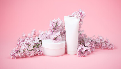 Face and body skin care. A set of cosmetic creams and balms in white tubes and cans on a coral background with sprigs of lilac flowers. Spa treatments for home care. Home rejuvenation and moisturizing