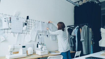Female clothing designer is taking sketches from studio table and putting them on wall with other...