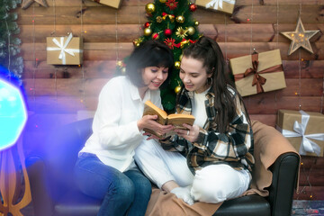 Mom and adult daughter are having fun reading a book on Christmas night against the background of a decorated Christmas tree.