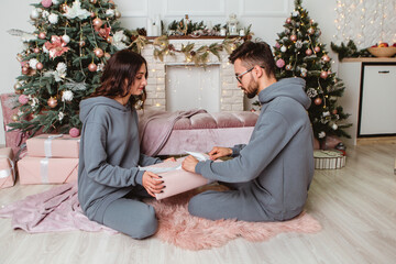 Lovely young romantic couple sitting open gifts presents garlands home cosy interior atmosphere New Year Christmas tree decorations holiday party celebrating concept winter evening 
