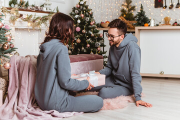 Lovely young romantic couple sitting open gifts presents garlands home cosy interior atmosphere New Year Christmas tree decorations holiday party celebrating concept winter evening 