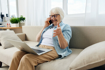 a joyful woman is sitting relaxed on a cozy sofa talking on a smartphone, holding a laptop on her lap while working from home, and gesticulating with her hand