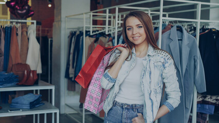 Portrait of happy and attractive girl standing in clothing shop with colourful bags, smiling gladly and looking at camera. Modern clothes is hanging in background.