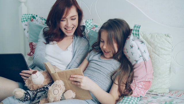 Little cute daughter with young loving mother watching funny pictures and laughing while reading fairy tale book sitting together in cozy bright bedroom at home