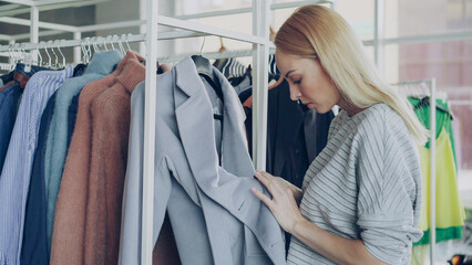 Close-up shot of young woman choosing coat in shop. First she is examining fabric carefully, looking at buttons, underlinimg and belt, then taking coat off hanger and taking it to fitting room