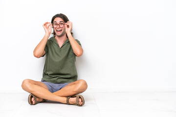 Young handsome man sitting on the floor isolated on white background with glasses and surprised