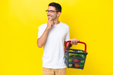 Young handsome man holding a shopping basket full of food over isolated yellow background looking...