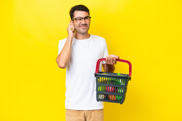 Young handsome man holding a shopping basket full of food over isolated yellow background...