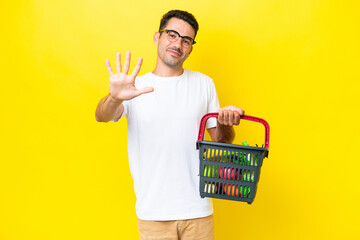 Young handsome man holding a shopping basket full of food over isolated yellow background counting...