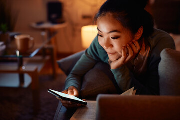 Fototapeta na wymiar Smiling Asian woman using smart phone while relaxing on sofa at home.