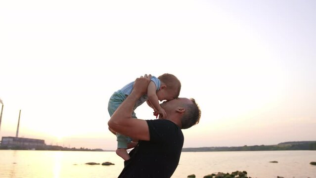 Loving dad talking to and kissing his little son. Laughing baby boy stepping his bare feet by father's chest. River at backdrop.