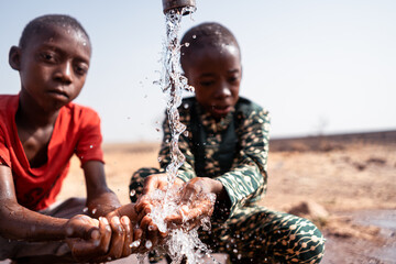 Small malnourished African boys at a village well refreshing themselves; concept of drought, famine and climate change - obrazy, fototapety, plakaty