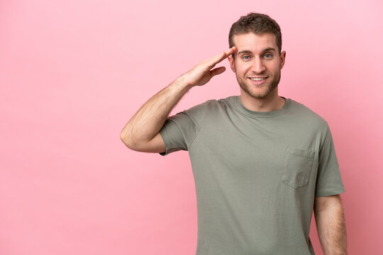 Young caucasian man isolated on pink background saluting with hand with happy expression