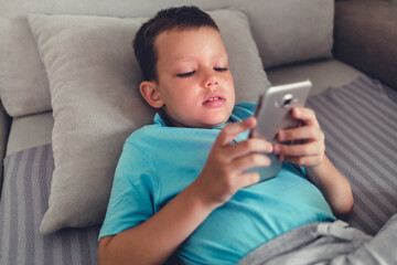 Smiling kid lying down on couch using smartphone. Boy using smart phone in the living room at home during the day.