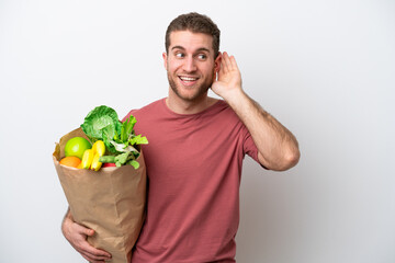 Young caucasian man holding a grocery shopping bag isolated on white background listening to something by putting hand on the ear