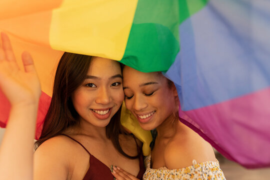 two multiracial lesbian women, of Hispanic and Asian origin, wrapped in a gay pride rainbow flag