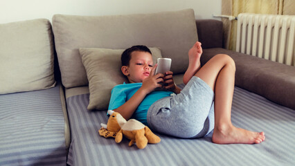 Cute boy playing with a mobile on sofa at home. Cute little kid focused on smartphone. Leisure, children, technology, internet communication and people concept.