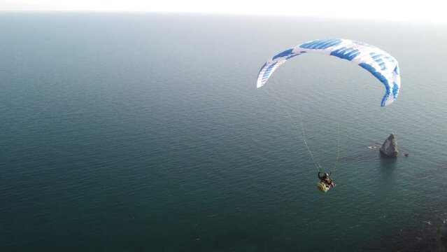 Aerial drone view of a man flying a white and blue paraglider over a hill and trees to the sea waves near the rocks. Active paraglider flight over the seascape with clear skies at suset. Extreme sport