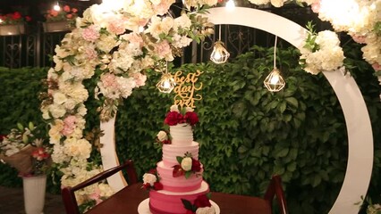 Wedding cake with best day ever sign on green garden decorated for wedding party at night. White and marsala creative decor with circle wedding arch decorated with roses and flowers, fairy lights.