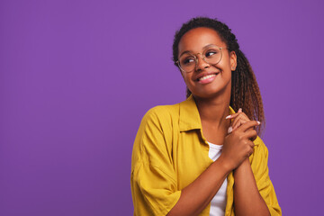 Young cheerful African American woman millennial look around and smile when see friend or great promotional offer dressed in casual oversize shirt stands on purple studio background
