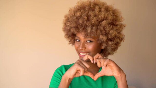 beautiful smiling dark-skinned woman in green t-shirt playfully and flirting shows air kiss and  heart shape with hands. Happy positive love romantic emotions.  Light beige background video footage