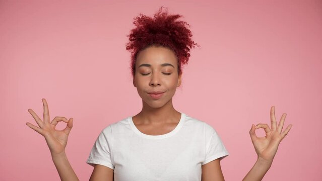 Close up African American redheaded curly woman hold spreading hands in yoga pose gesture. Smiling African female breathes deeply relax meditate try to calm down on isolated pink background