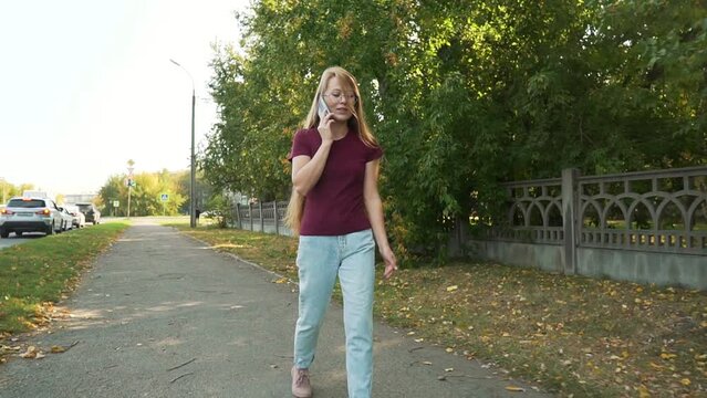 Positive casual woman talking smartphone in an urban street. Smiling girl calling mobile phone outside. Lady walking down the road in a city.