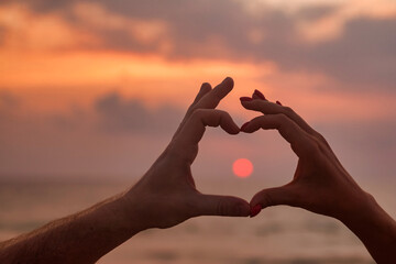 Silhouette hands of couple are lovely heart shape with sunset on through hand. Woman and man hands in heart shape with sunrise in middle and sea (ocean) background. Togetherness and love. Copy space