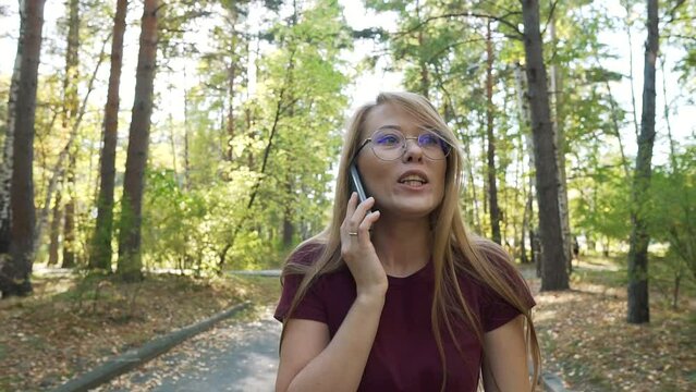 Excited woman talking smartphone in a park, telling news or gossip. Thrilled girl chatting mobile phone outside. Lady walking down the path in a forest. Zoom out.