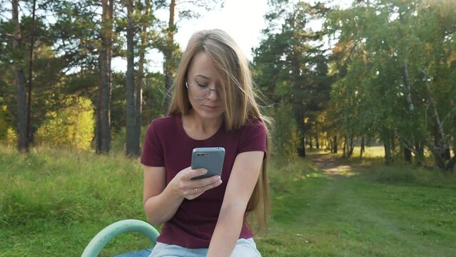 Calm woman using smartphone in a park, reading online news or media, typing sms. Girl chatting in mobile phone messengers outside, sitting in a park. Pan shot.
